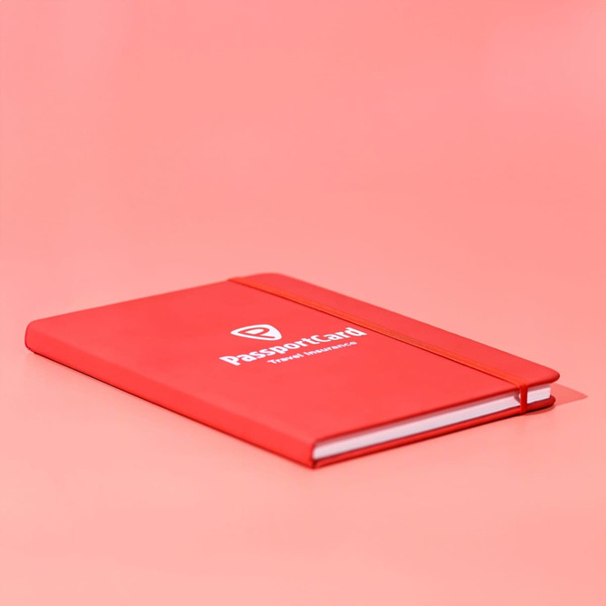 A red A5 notebook with a logo on the cover and an elastic band closure, set against a pink background.