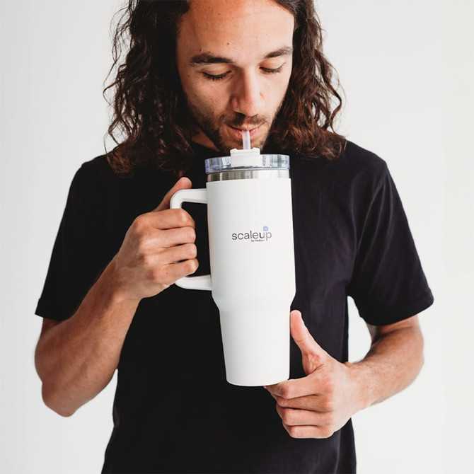 A drink bottle in white with a transparent lid and straw, featuring a logo, held by a person.