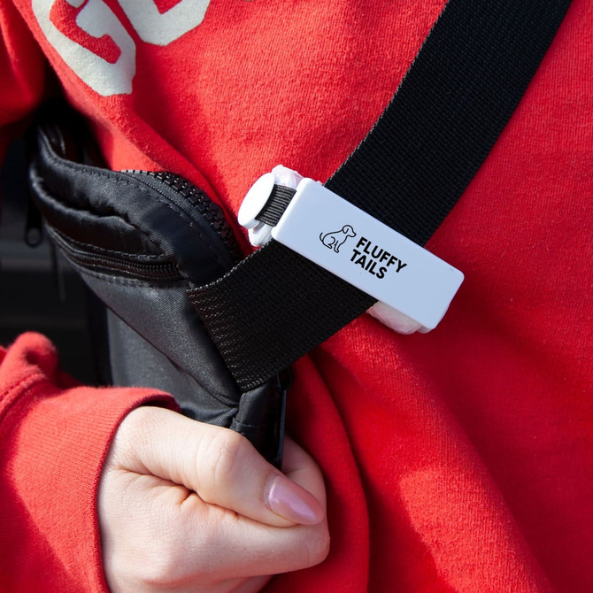 A black pet bag dispenser attached to a strap, featuring a white clip with a logo and a person's hand wearing a red sweatshirt.