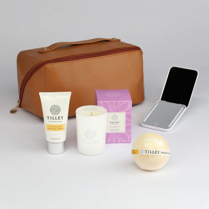 A tan toiletry bag accompanied by a skincare tube, candle, bath bomb, and a compact mirror, all branded with Tilley.