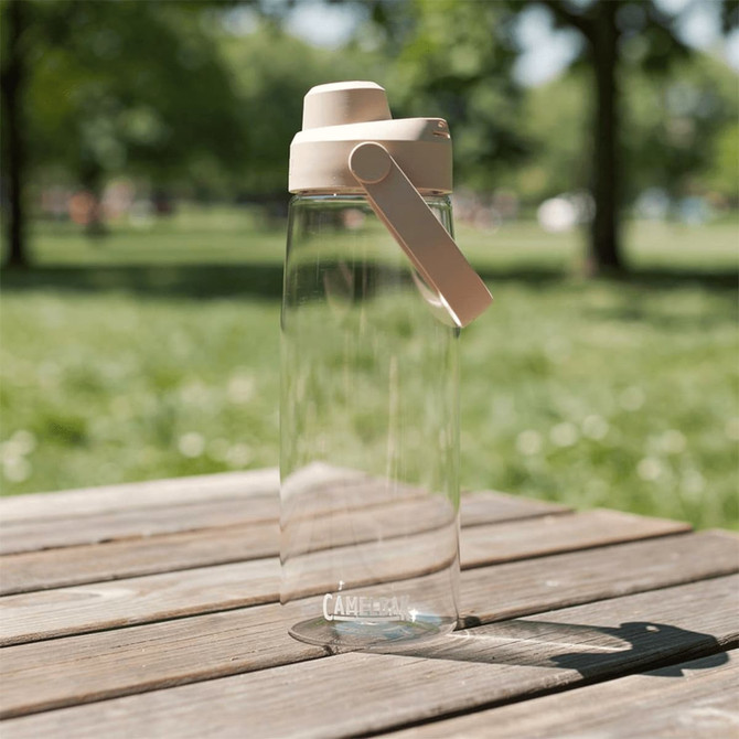 A drink bottle with a clear body and a beige lid, featuring a handle, set on a wooden table in a grassy area.