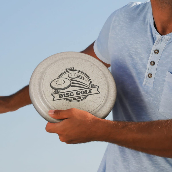A person holds a large grey disc with a disc golf design and logo, wearing a light blue shirt.