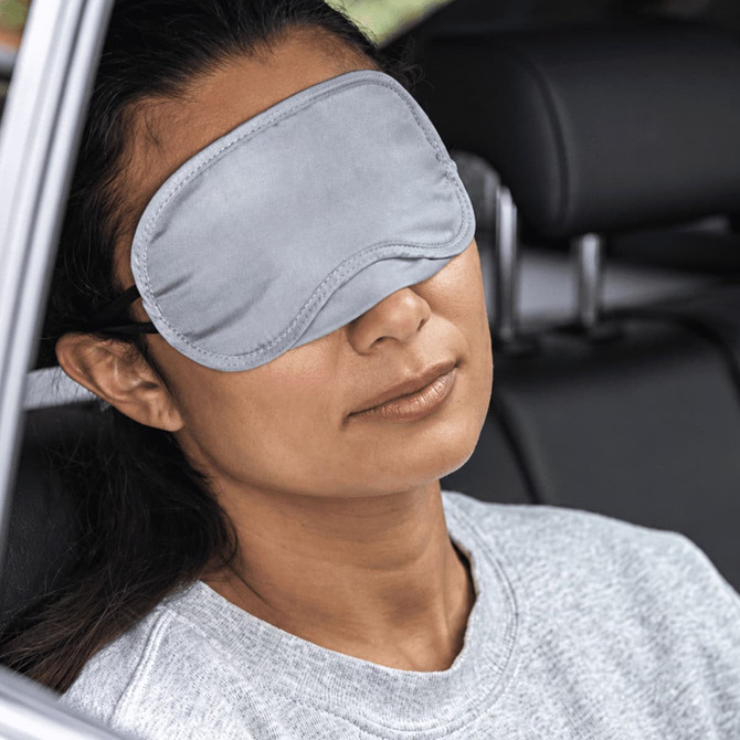 A grey eye mask rests over a woman's eyes as she relaxes in a car seat, featuring a logo on the mask.