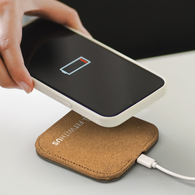 A square wireless charger in a beige cork finish with a smartphone placed on top, displaying a low battery icon.
