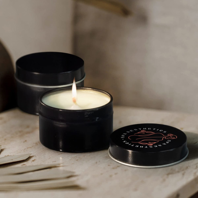 A black travel candle with a lit wick, placed beside its matching lid on a neutral surface. It features a logo on the lid.