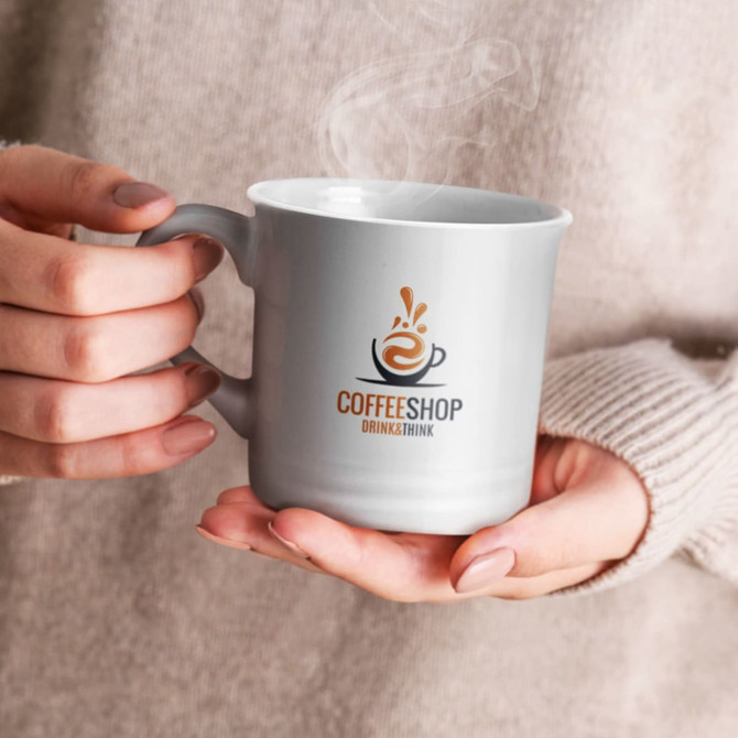 A grey coffee mug with a logo, being held by a person wearing a beige sweater, with steam rising from the mug.