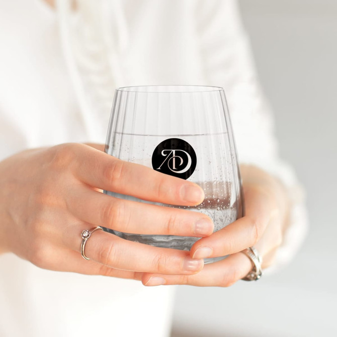 A stemless low ball glass in clear, held by a person, featuring a logo on the front.