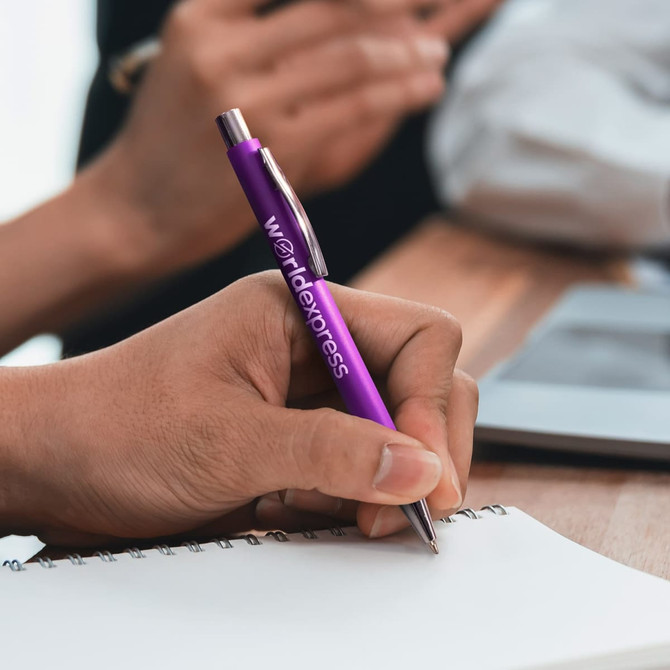 A purple plastic pen with a silver tip, held in hand, writing on a notebook.
