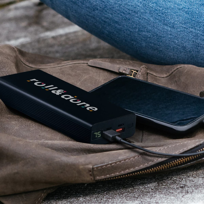 A black power bank with a logo connected to a smartphone, resting on a brown bag.