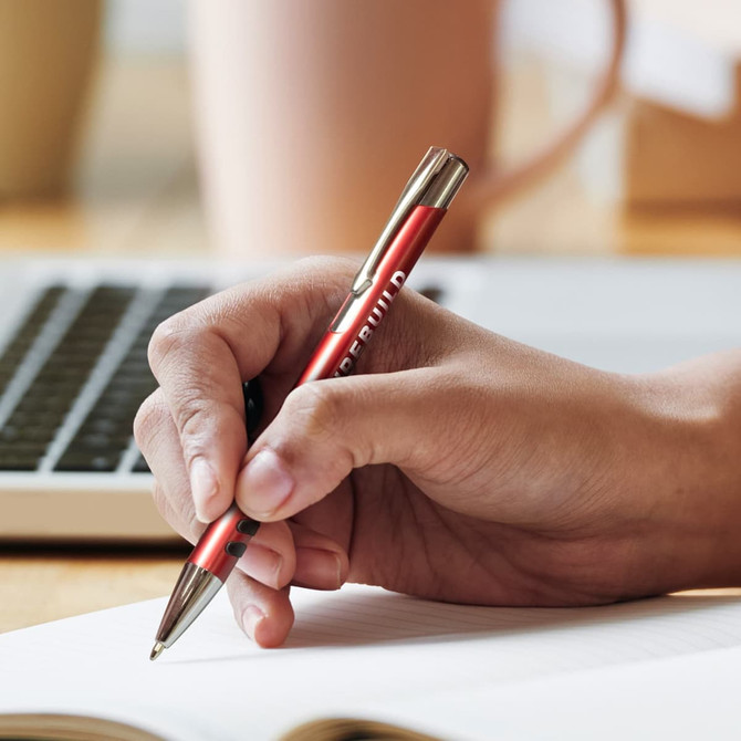 A metal pen with a red barrel and silver accents held over a notebook, with a laptop in the background.