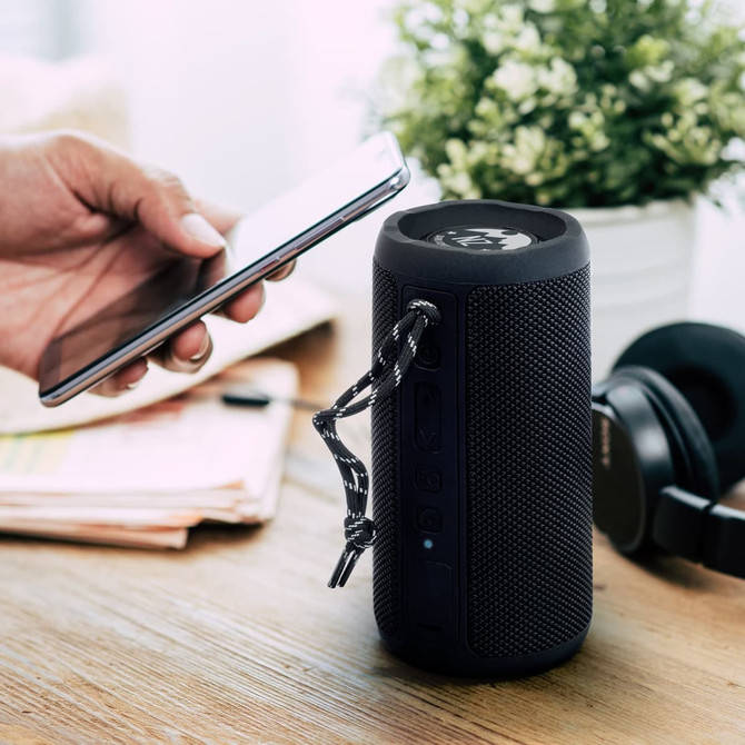A black Bluetooth speaker with textured surface, surrounded by a phone, headphones, and a small plant on a wooden table.