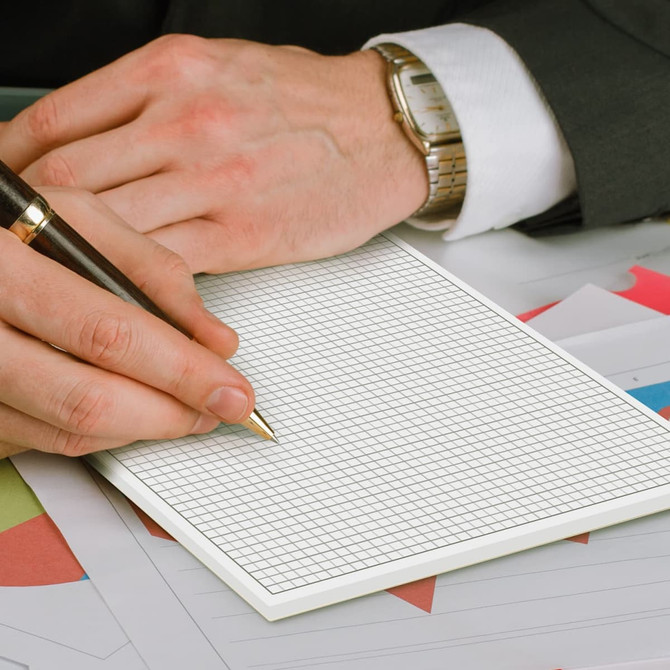 A hand holding a pen writes on a blank, gridded A4 notepad with a textured surface. The background features colourful paper.