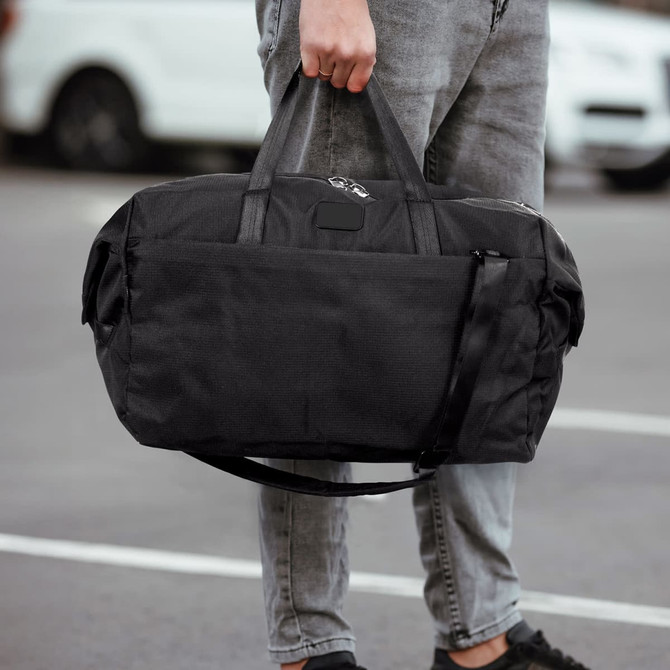 A black duffle bag is being held by a person, featuring dual handles and a shoulder strap.