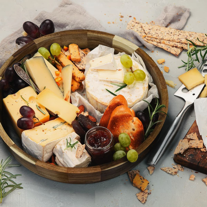 A wooden platter tray filled with various cheeses, grapes, crackers, and a jar of jam, styled with herbs.