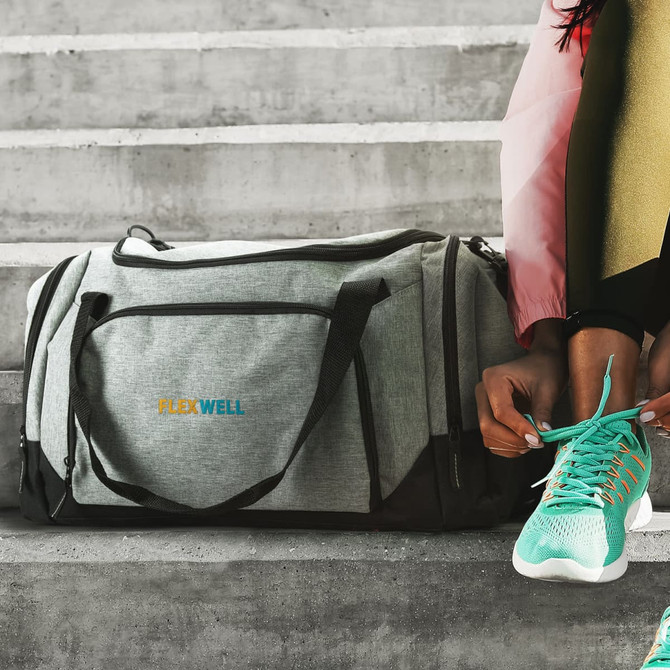 A grey and black duffle bag sits on a concrete surface, with a logo, beside a person tying their shoe.