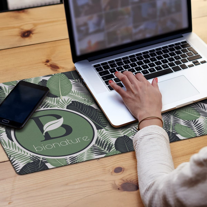 A desk mat featuring a leaf pattern in green and white, with a logo design, beside a laptop and smartphone.