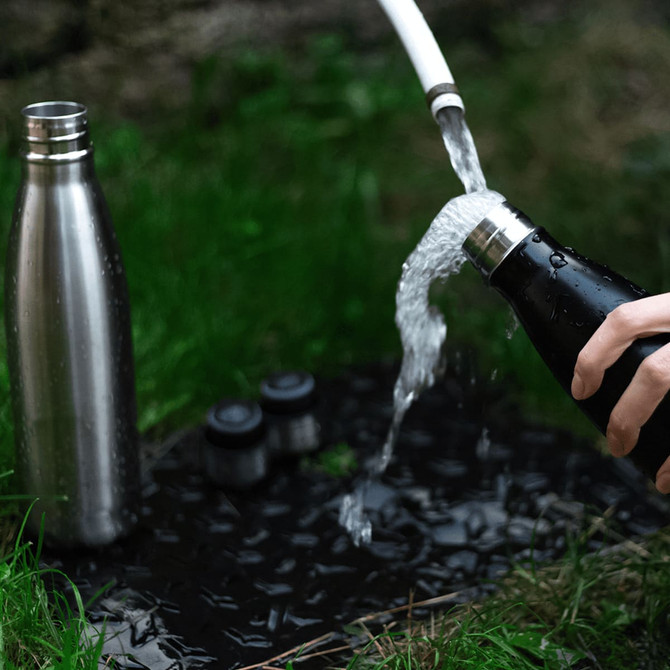 A drink bottle in stainless steel and black, with water being poured from it outdoors on a grassy surface.