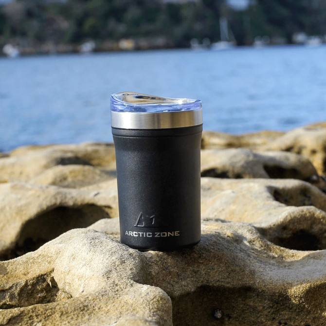 A drink bottle in black with a silver lid, featuring branding, set against a rocky background near water.