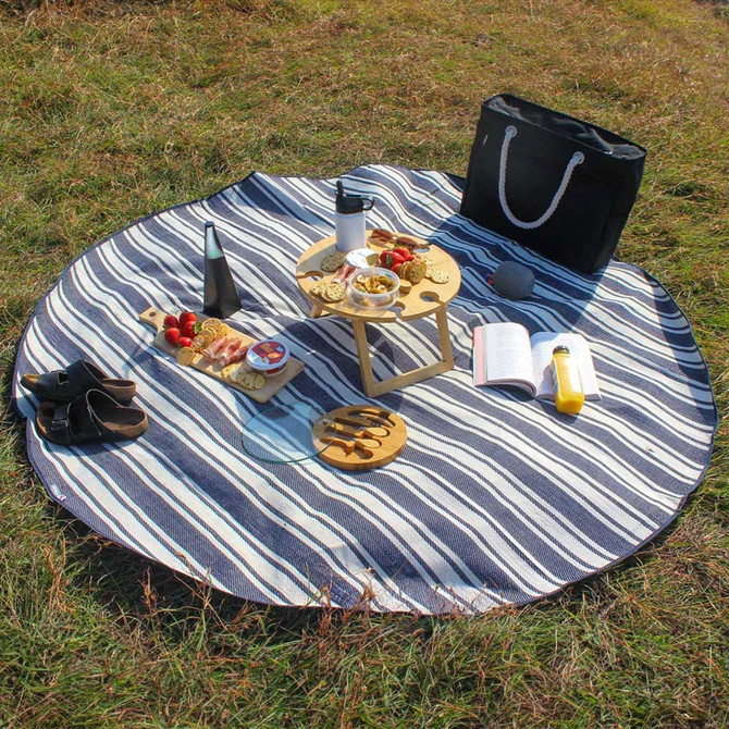A round picnic rug with navy and white stripes, surrounded by food, drinks, and accessories on grass.