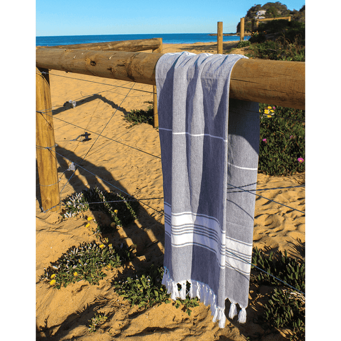 A grey and white striped Turkish towel draped over a wooden fence on a sandy beach, with ocean in the background.