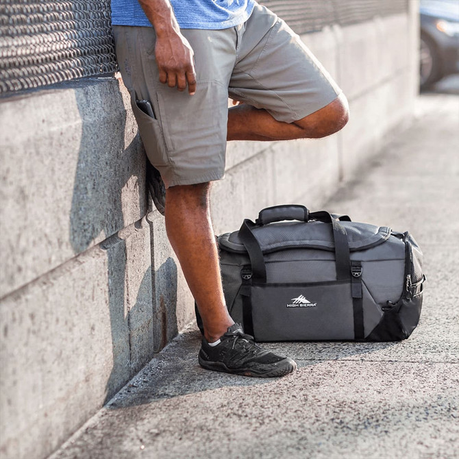 A duffel backpack in grey with black accents, resting on the ground beside a person in shorts and sneakers.