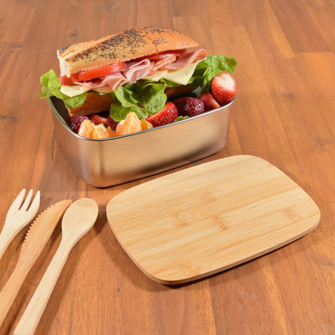 A metal lunch box filled with a sandwich, lettuce, strawberries, and a cutting board made of wood beside it.