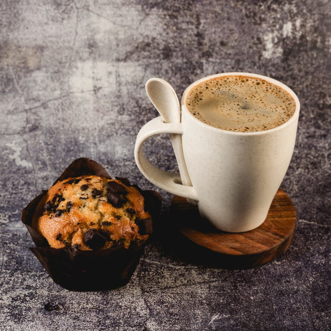 A white wheat fibre cup with coffee, accompanied by a chocolate chip muffin on a wooden coaster.