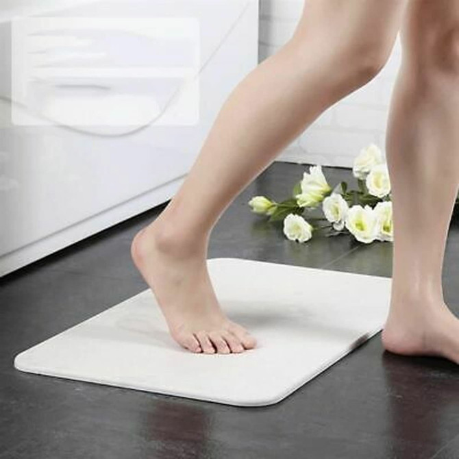 A soft white bath mat with a foot stepping onto it, placed on a tiled floor near flowers. It has a logo.