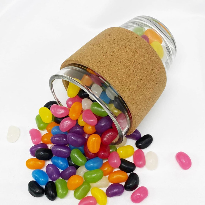 A glass coffee cup with a cork band, filled with colourful jelly beans, some spilling onto a white surface.