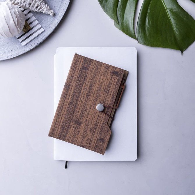 A wooden-textured notebook with a button closure rests on a white notepad, surrounded by a decorative plate and greenery.