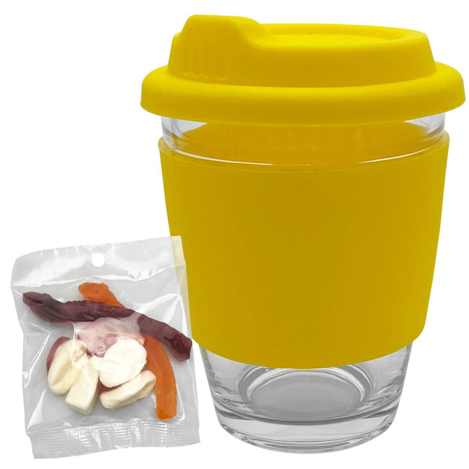 A reusable glass coffee cup with a yellow silicone lid and sleeve, accompanied by a clear bag of assorted jelly sweets.