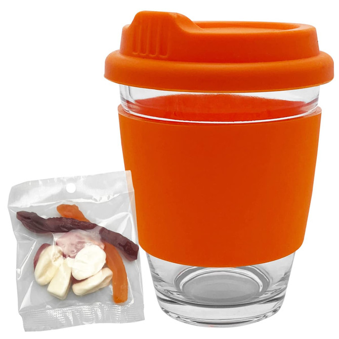 A reusable coffee cup in clear glass with an orange silicone sleeve and lid, accompanied by a bag of jelly candies.