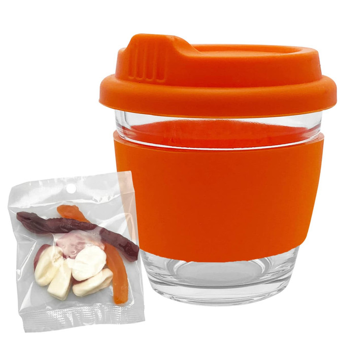A clear glass coffee cup with an orange silicone lid and sleeve, accompanied by a bag of mixed jelly lollies.