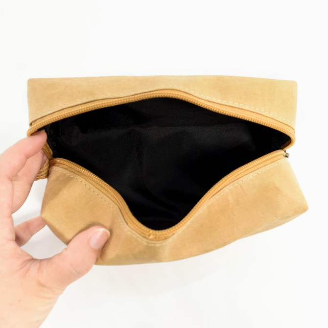A brown kraft paper toiletry bag with a black interior and a zip closure, held open by a hand.