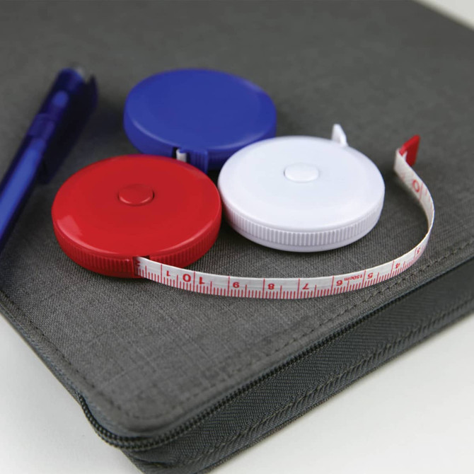 A compact tape measure in red, blue, and white, lying next to a pen on a grey surface. It has a logo.
