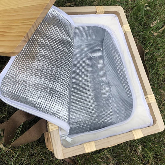 A square wicker picnic cooler basket with a wooden lid and a silver insulated interior, resting on green grass.