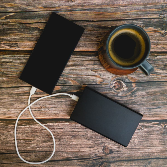 Two black power banks with white charging cables, placed alongside a cup of coffee on a wooden surface.