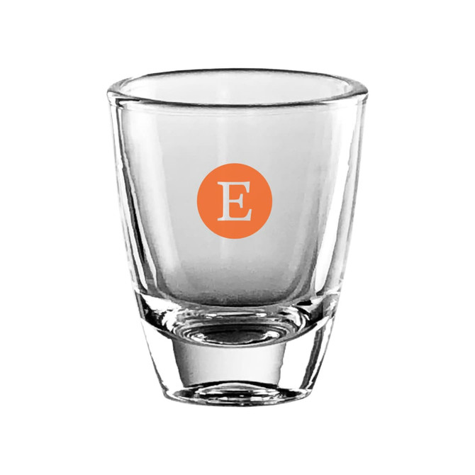 A clear glass shot glass with a solid base and an orange circular logo containing the letter "E."