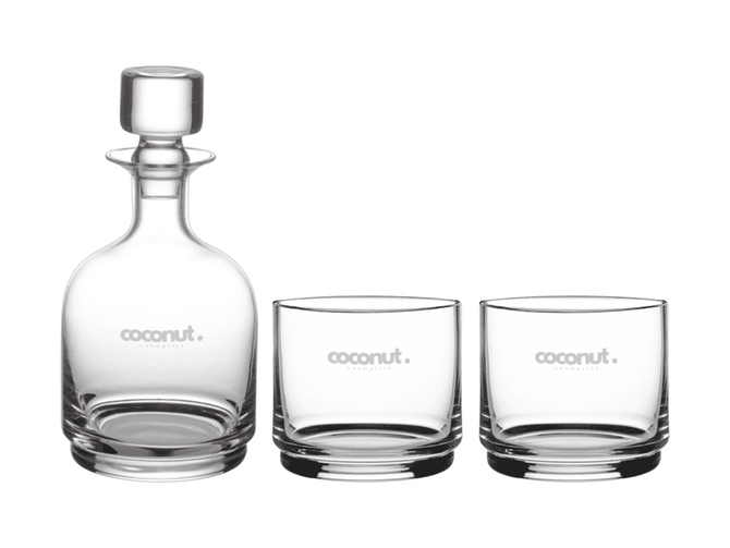 A glass decanter and two matching tumblers, all clear with a subtle logo.