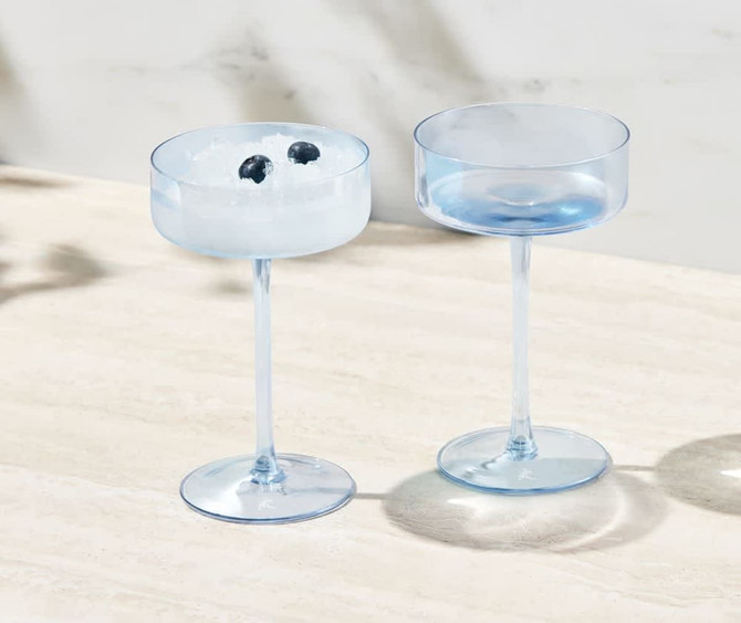Two clear blue coupe glasses, one filled with ice and blueberries, elegantly presented on a light surface.
