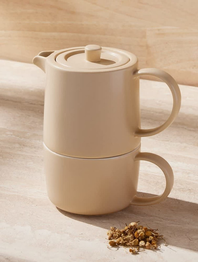 A beige tea set with a teapot and cup stacked together, accompanied by dried flowers, on a light stone surface.
