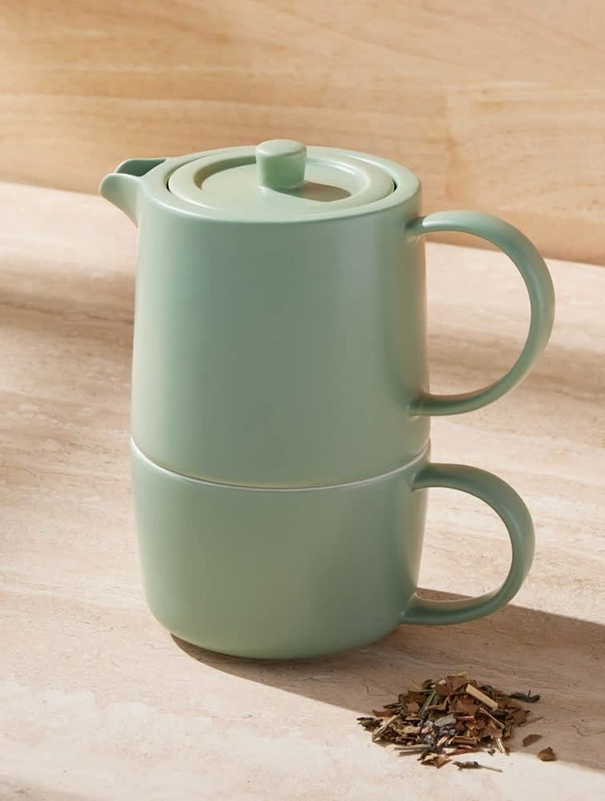 A green tea infuser set with a stacked teapot and cup, accompanied by loose tea leaves on a surface.