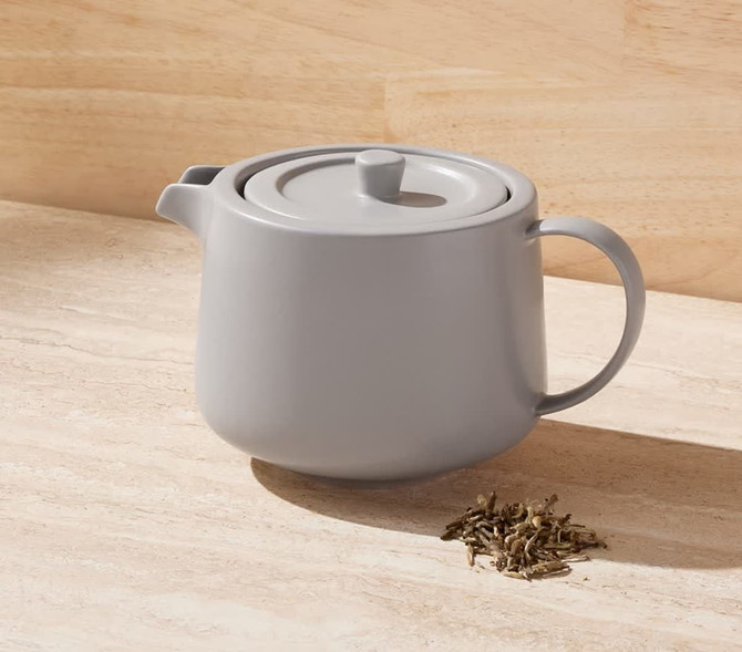 A grey teapot with infuser, placed on a light stone surface, accompanied by loose tea leaves.