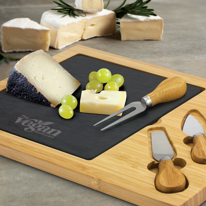 A slate cheese board featuring assorted cheeses, grapes, and stainless steel knives with wooden handles.