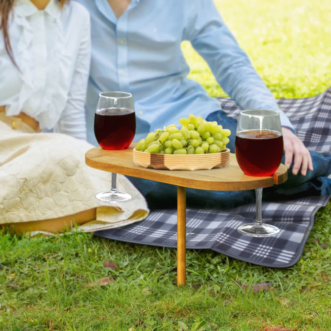 A wooden picnic serving board holds grapes, with two glasses of red drink on a green lawn, accompanied by a checkered blanket.