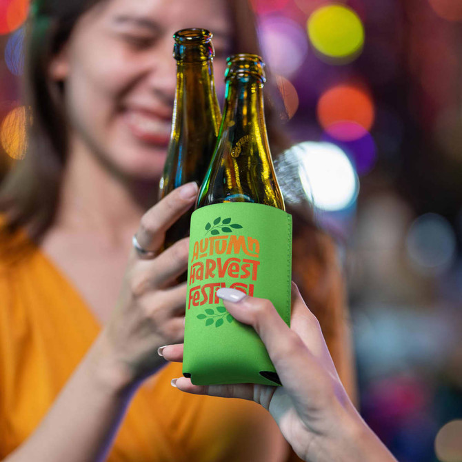 A drink bottle cooler in green featuring a logo design, held by two hands against a blurred, colourful background.