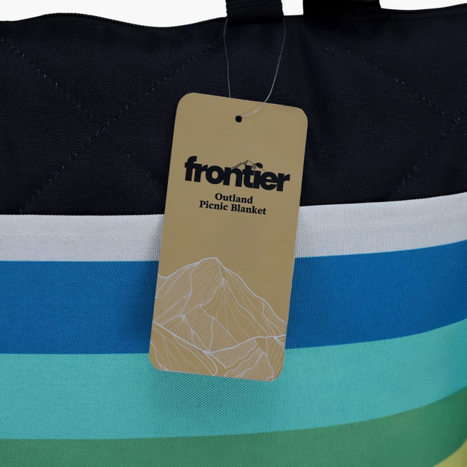 A striped picnic blanket in shades of blue, green, and white, featuring a hang tag with a logo.