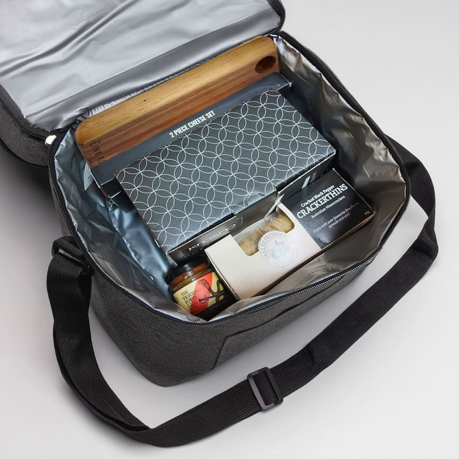A cooler bag in grey containing a cheese kit, crackers, a jar, and a wooden board. It has a logo inside.