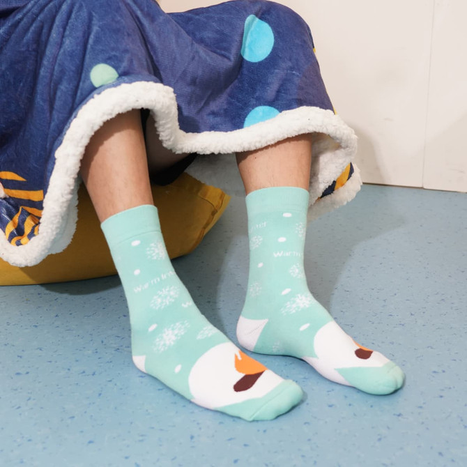 A pair of mint green socks featuring a winter design with snowflakes and a character, worn with a cosy blanket.