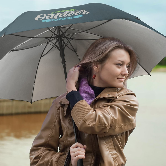 A black and grey umbrella is held by a woman with long hair, wearing a brown jacket, near a body of water.